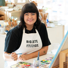 Load image into Gallery viewer, Person wearing a 'Big Bite Studios' apron, sitting at a table with art supplies.