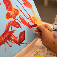 Load image into Gallery viewer, Person painting red lobsters on a blue canvas with a paintbrush.
