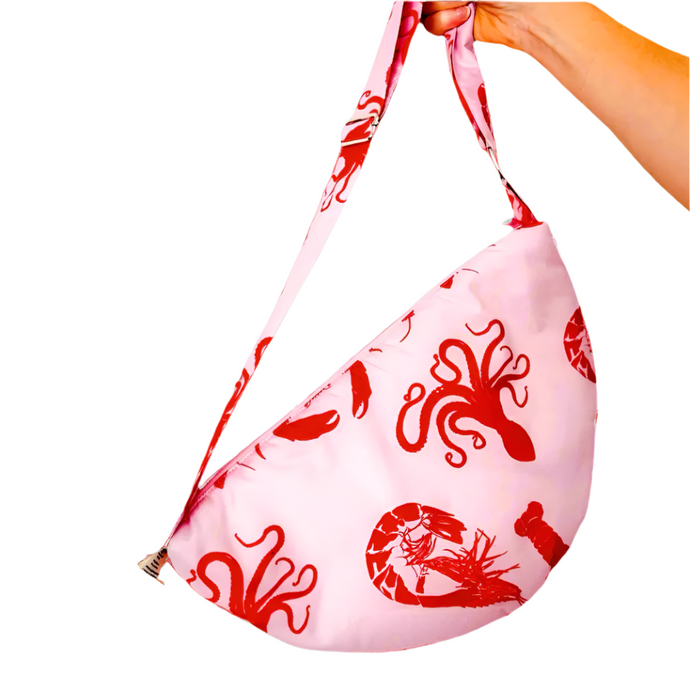 product photo of fabric sling style bag. Pastel pink bag with red octopus and shrimp patterns. Hand is holding bag by the long strap