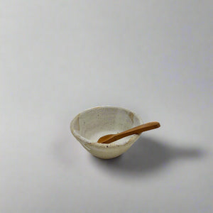 Product photo of a miniature wooden spoon, sitting in a small ceramic bowl resting against the edge. 