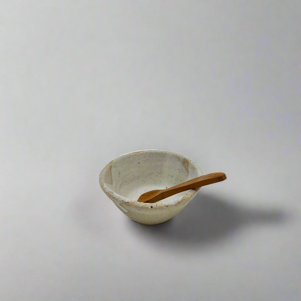 Product photo of a miniature wooden spoon, sitting in a small ceramic bowl resting against the edge. 